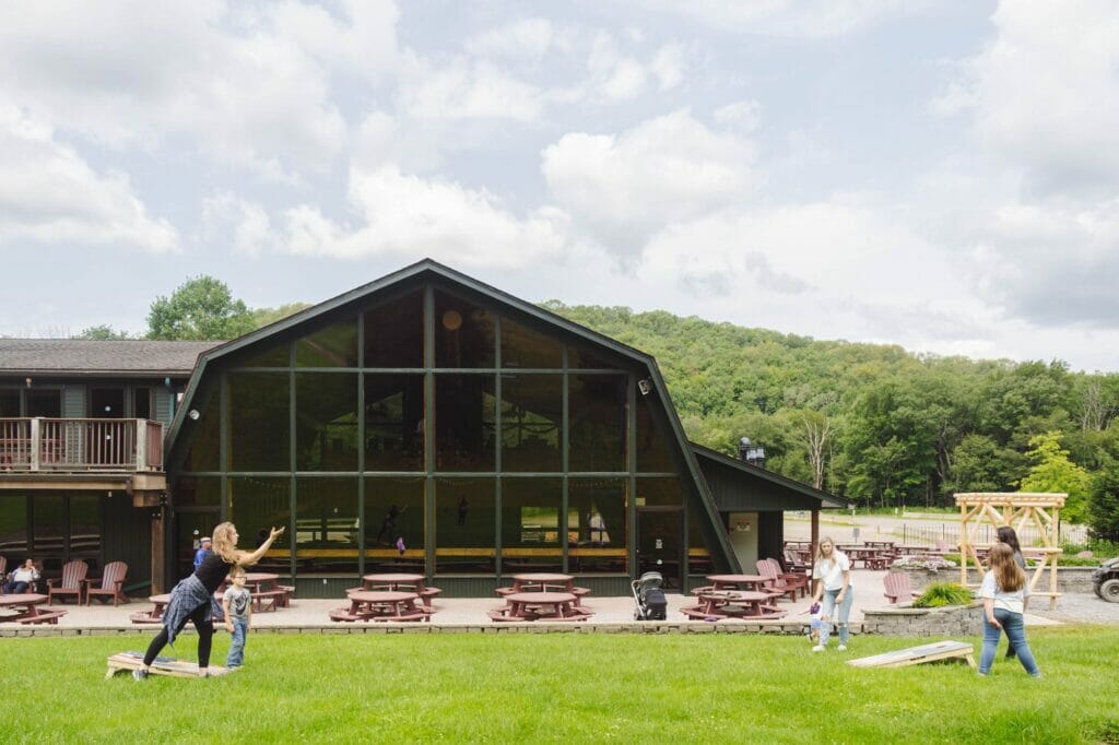 A woman plays a lawn game with a child in front of a rustic building with large windows, surrounded by picnic tables and greenery, under a cloudy sky and forested hills.