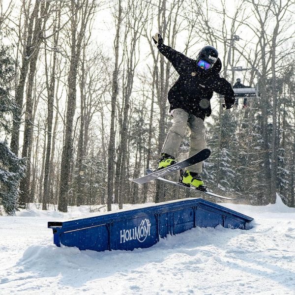 Snowboarder performing a jump off a blue ramp with "HOLIMONT" text, surrounded by snowy trees and a wintry forest environment.