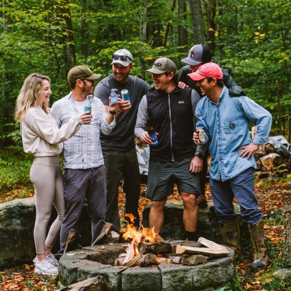 A group of six people stand around a campfire, holding drinks and smiling, in a wooded area surrounded by autumn leaves and logs.