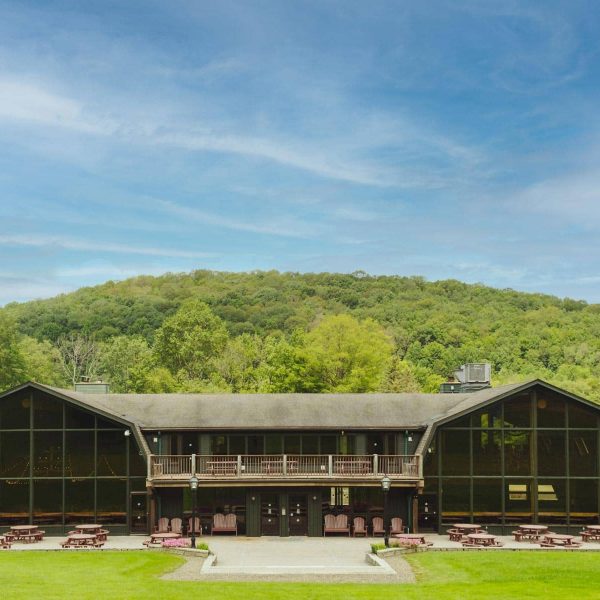 A large A-frame building features a glass facade, overlooking picnic tables and neatly mown grass, set against a backdrop of lush green hills under a clear blue sky.